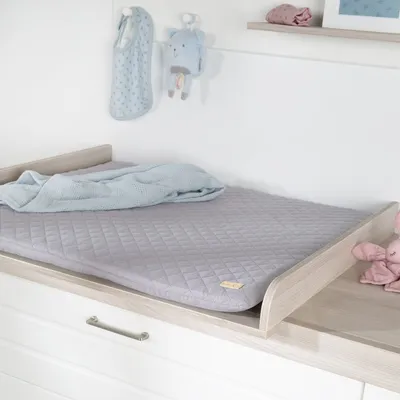 A baby changing mat with a blanket on it, placed on top of a dresser. There are baby toys hanging on the wall behind it.