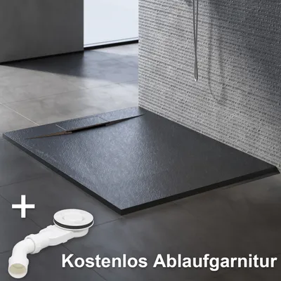 A black shower tray with a slate-like texture is shown on a tiled floor, with a white drain assembly in the foreground. Text indicates a free drain set.