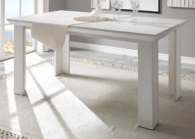 A white dining table with a runner and glasses on it, set in a bright room with sunlight streaming through a window.
