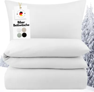 A stack of white bedding, including a pillow and folded sheets, with a tag attached to the pillow. A snowy forest scene is visible in the background.