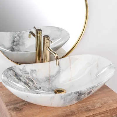 A marble basin with gold faucets is shown on a wooden surface, with a round mirror in the background.