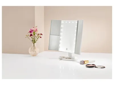 A makeup mirror with LED lights and two side mirrors is displayed on a white surface, with a vase of flowers to the left and makeup brushes and a compact to the right.