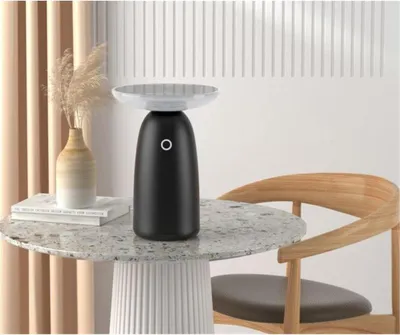 A black solar table lamp with a white top sits on a speckled round table next to a vase with dried grass and books, with a wooden chair in the background.