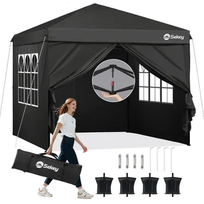 A woman is walking with a folded tent bag in front of an open black pop-up tent. The tent has windows and is set up outdoors on a white surface.