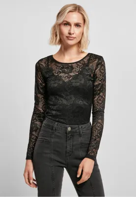 A woman is posing in front of a plain background, wearing a black lace long-sleeved top and dark wash jeans.