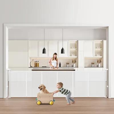 A child is pushing a dog in a wooden cart in front of a baby gate, with a woman in the background in a kitchen.