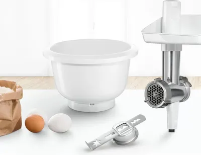 A collection of kitchen appliance attachments, including a mixing bowl, a meat grinder, and a whisk attachment, are displayed on a white surface with a light gray background.