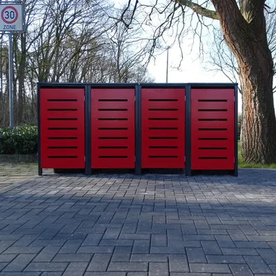 Four red trash bins with black frames are lined up on a paved area, with trees and a street sign in the background.