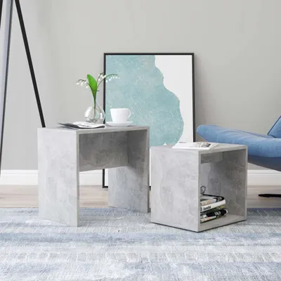 Two cube-shaped side tables with a concrete-like finish are placed on a blue and white striped rug in a living room setting. A vase with flowers and a cup of coffee sit on the left table, while books are stacked on the right table. A blue chair and a frame