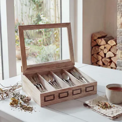 A wooden cutlery box with a glass lid is placed on a white table, filled with cutlery. In the background, a window shows a garden, and a stack of firewood is visible.