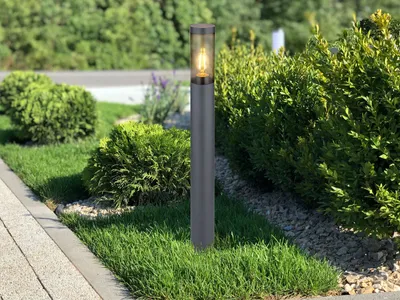 A modern outdoor lamp post stands in a garden setting with manicured bushes and grass.