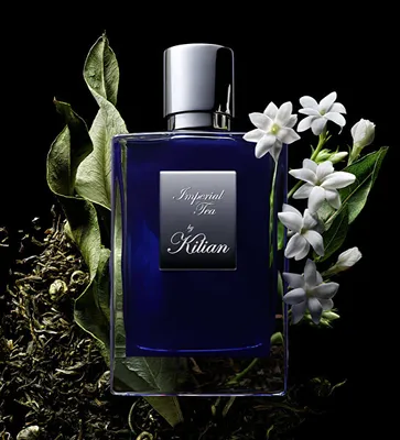 A bottle of Kilian Imperial Tea perfume is displayed with dried tea leaves and jasmine flowers against a black background.