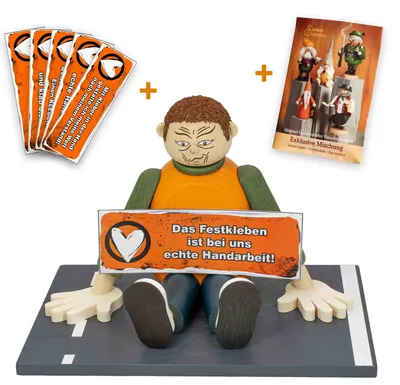 Ein Holzfigur sitzt auf einer Straße und hält ein Schild mit der Aufschrift 'Das Festkleben ist bei uns echte Handarbeit' während daneben mehrere Karten und eine Packung mit Figuren zu sehen sind