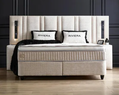 A luxurious beige boxspring bed with a tall, upholstered headboard and two decorative pillows.