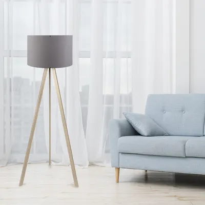 A floor lamp with a gray shade and a wooden tripod base stands next to a light blue sofa in a room with sheer white curtains.