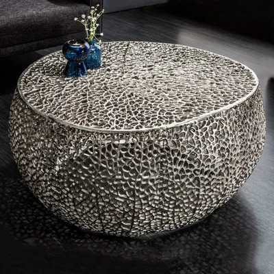 A round, silver coffee table with an intricate, organic design sits on a dark floor, with a dark gray sofa visible in the background. Two small blue vases with delicate white flowers are placed on the table.