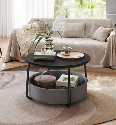 A modern living room scene featuring a round black coffee table with a fabric basket underneath, placed on a patterned rug in front of a sofa.