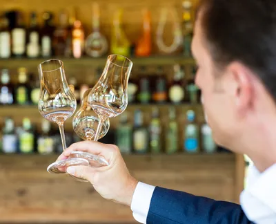 A person holding three rum glasses in front of a bar with bottles on shelves.