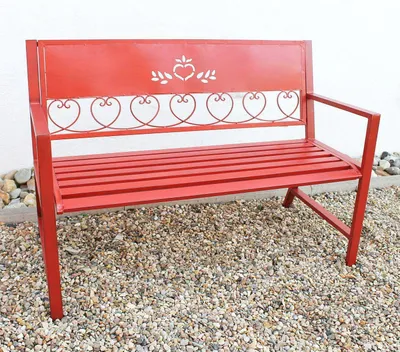 A red metal garden bench with heart-shaped decorative elements on a gravel surface in front of a white wall.
