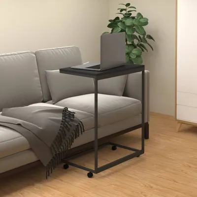 A laptop sits on a black C-shaped side table next to a sofa with a gray throw blanket.