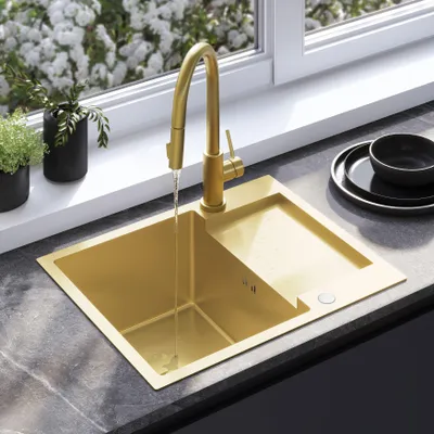 A gold sink with a faucet running water, set on a dark countertop with plants in the background and a window above.
