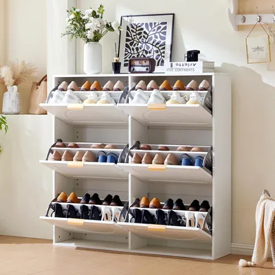 A white shoe cabinet with multiple shelves and flip-down doors, filled with shoes, placed in a room with a plant and artwork in the background.