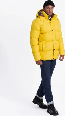 A man wearing a yellow puffer jacket and blue jeans, posing against a white background.
