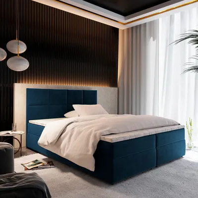 A bedroom scene featuring a blue boxspring bed with white bedding, set against a dark, textured wall and a window with sheer curtains.