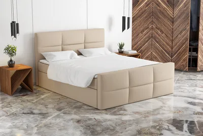 A beige upholstered box spring bed with white bedding is shown in a modern bedroom setting with a wooden wardrobe in the background.