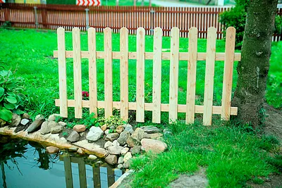 A small wooden fence is placed in a garden next to a pond and a tree. The fence is made of light-colored wood and has a straight design. The background shows a larger wooden fence and green grass.