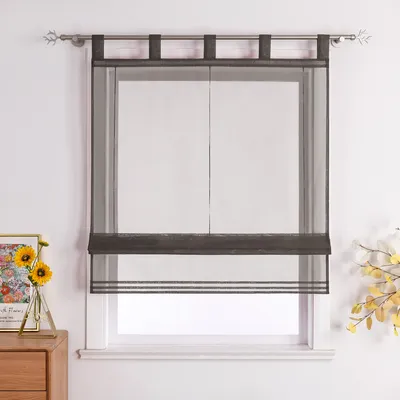 A dark gray sheer Roman shade with tabs hangs on a window, with a vase of sunflowers and a framed picture on a wooden dresser to the left.