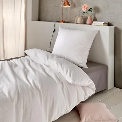 A neatly made bed with white bedding and a pink pillow, next to a bedside table with a lamp and flowers.