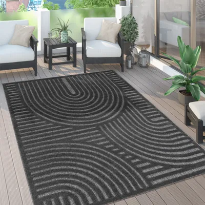 An outdoor rug with a geometric pattern is placed on a wooden deck, with outdoor furniture and plants visible.