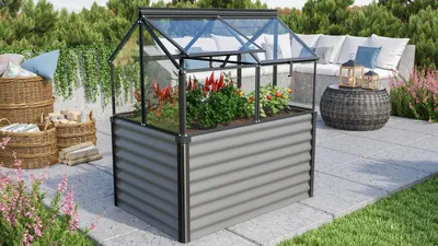 A raised garden bed with a greenhouse top, filled with plants, sits on a paved patio next to outdoor furniture and baskets.