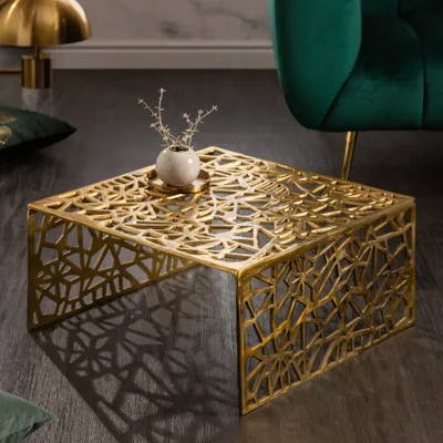 A gold abstract coffee table with a small vase on top, placed on a dark floor next to a green velvet armchair.