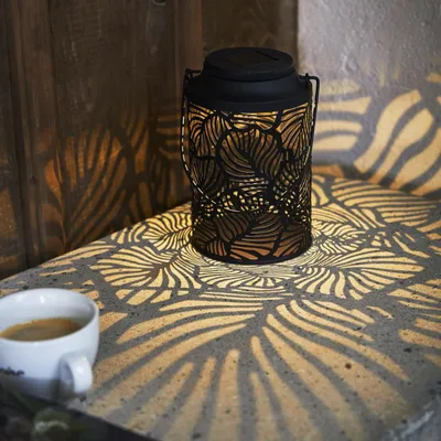 A black metal lantern with a leaf pattern is placed on a stone surface next to a cup of coffee. The lantern casts intricate leaf-shaped shadows on the surface and the wooden wall behind it.