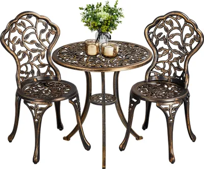 A bronze bistro set with two chairs and a table, decorated with a leaf motif, is displayed against a white background. A vase with greenery and two candle holders are placed on the table.