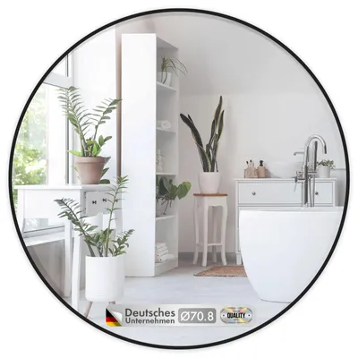 A round mirror with a black frame is shown in a bathroom setting, reflecting plants, shelves, and a bathtub.