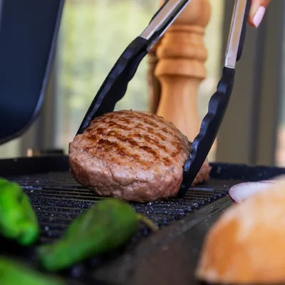A hamburger patty is being grilled on a grill, with green peppers and a bun in the foreground.