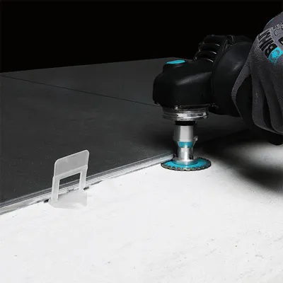 A hand wearing a glove is using a power tool to cut a tile. A tile leveling spacer is visible.