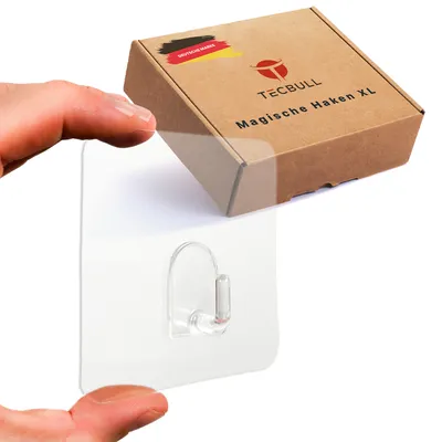 A hand is holding a transparent adhesive hook, with a product box for TECBULL Magische Haken XL in the background.