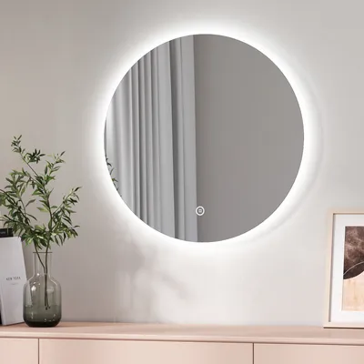 A round LED mirror with a touch switch is mounted on a wall above a light pink dresser. A vase with green leaves is placed on the dresser to the left of the mirror. A framed picture is on the right.