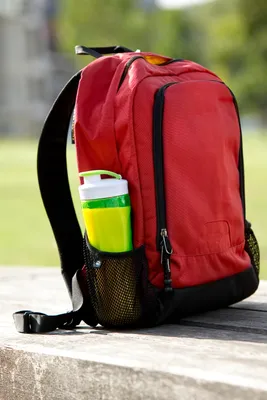 A red backpack with a green water bottle in the side pocket, resting on a wooden surface outdoors.