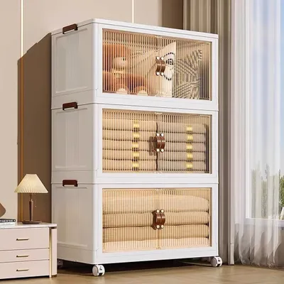 A tall, white, stackable storage unit with transparent doors and brown handles, filled with folded items and a teddy bear. It is placed next to a small chest of drawers with a lamp on it.