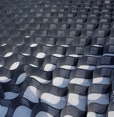A close-up, angled view of a black geogrid or honeycomb-like structure laid out on a light gray surface. The structure is made of interconnected cells with small perforations.