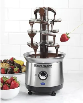 A chocolate fountain with chocolate flowing down the tiers, with strawberries and other fruits in the background.