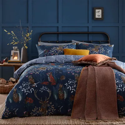 A cozy bedroom scene featuring a bed with a forest-themed duvet cover, pillows, and a throw blanket. The room has dark blue paneled walls and a small wooden table with a lantern and logs.