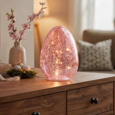 An Easter-themed decorative egg lamp with lights inside, placed on a wooden table next to a vase with flowers and a small nest with eggs.