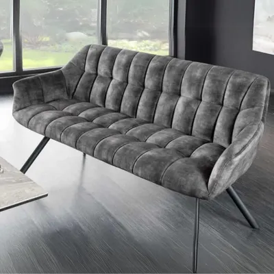 A modern gray sofa with tufted upholstery and black metal legs is placed in a room with large windows and a dark floor.