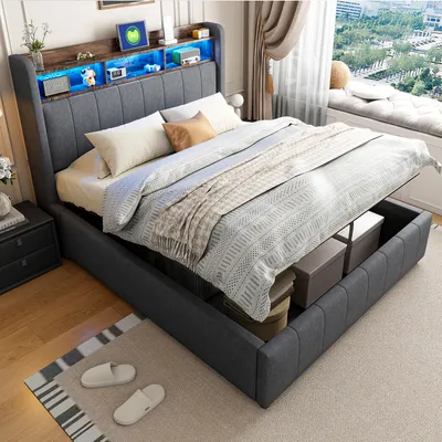 A bedroom scene featuring a modern upholstered bed with storage, illuminated by LED lights in the headboard. The bed is made with blankets and pillows, and the storage compartment underneath is partially open, revealing storage boxes.